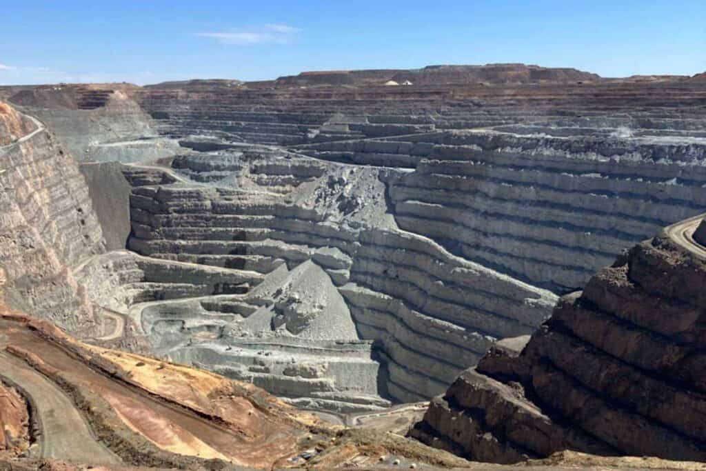 A vast open-pit mine known as the Superpit in Kalgoorlie Boulder, with intricate terraces descending into the earth, showcasing the scale of human engineering in resource extraction. The layered geological strata are visible, highlighting the depth and complexity of the mining operations under a wide blue sky.